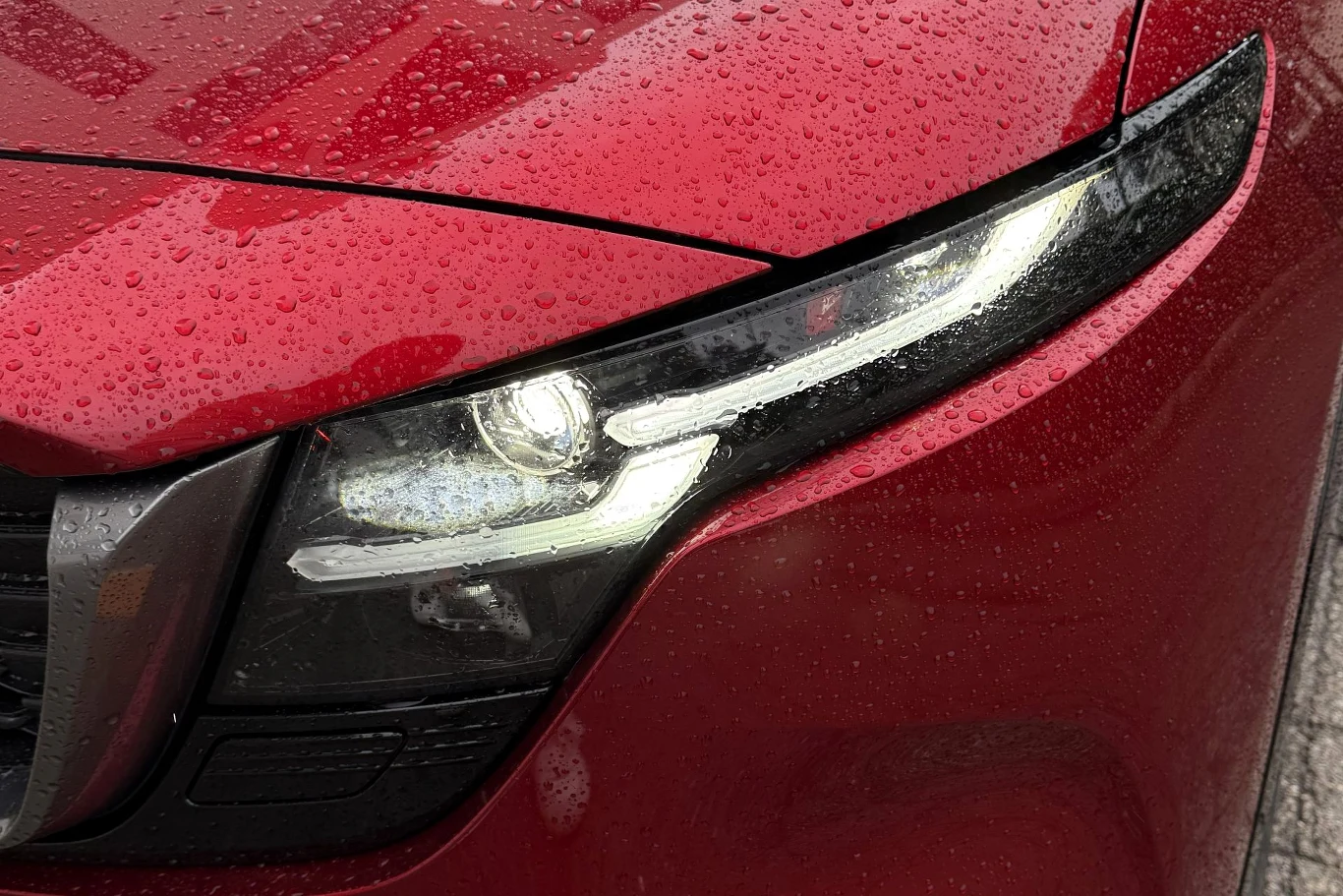 Zbliżenie na nowoczesny, wąski reflektor LED w czerwonym samochodzie pokrytym kroplami deszczu.