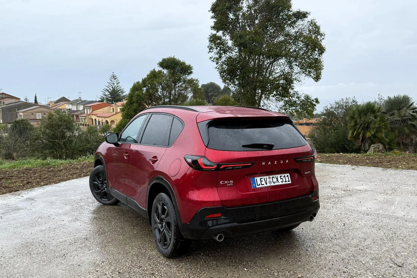 Czerwony SUV marki Mazda CX-5 z czarnymi felgami stoi na mokrej nawierzchni, w tle widać rzadkie drzewa oraz zabudowania mieszkalne.