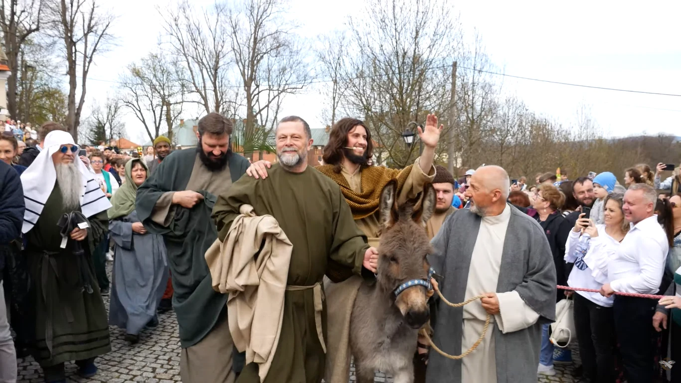 Grupa mężczyzn ubranych w stroje przypominające czasy biblijne prowadzi osła przez tłum ludzi, jedna z centralnych postaci z szerokim uśmiechem unosi rękę w geście powitania, wokół nich liczni obserwatorzy przyglądają się scenie.