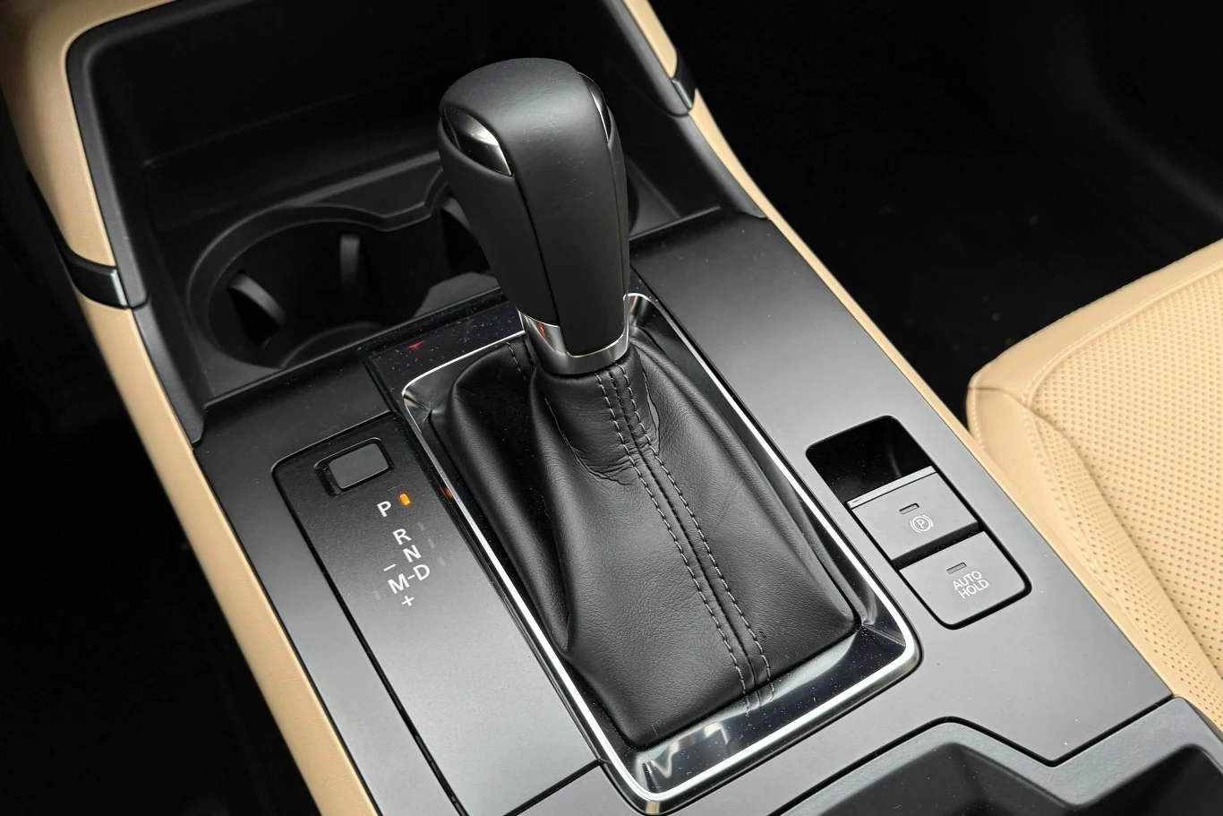 Czarna dźwignia automatycznej skrzyni biegów otoczona połyskującym panelem, widoczne oznaczenia pozycji biegów oraz przyciski funkcyjne obok podłokietnika i jasnego siedzenia pasażera.