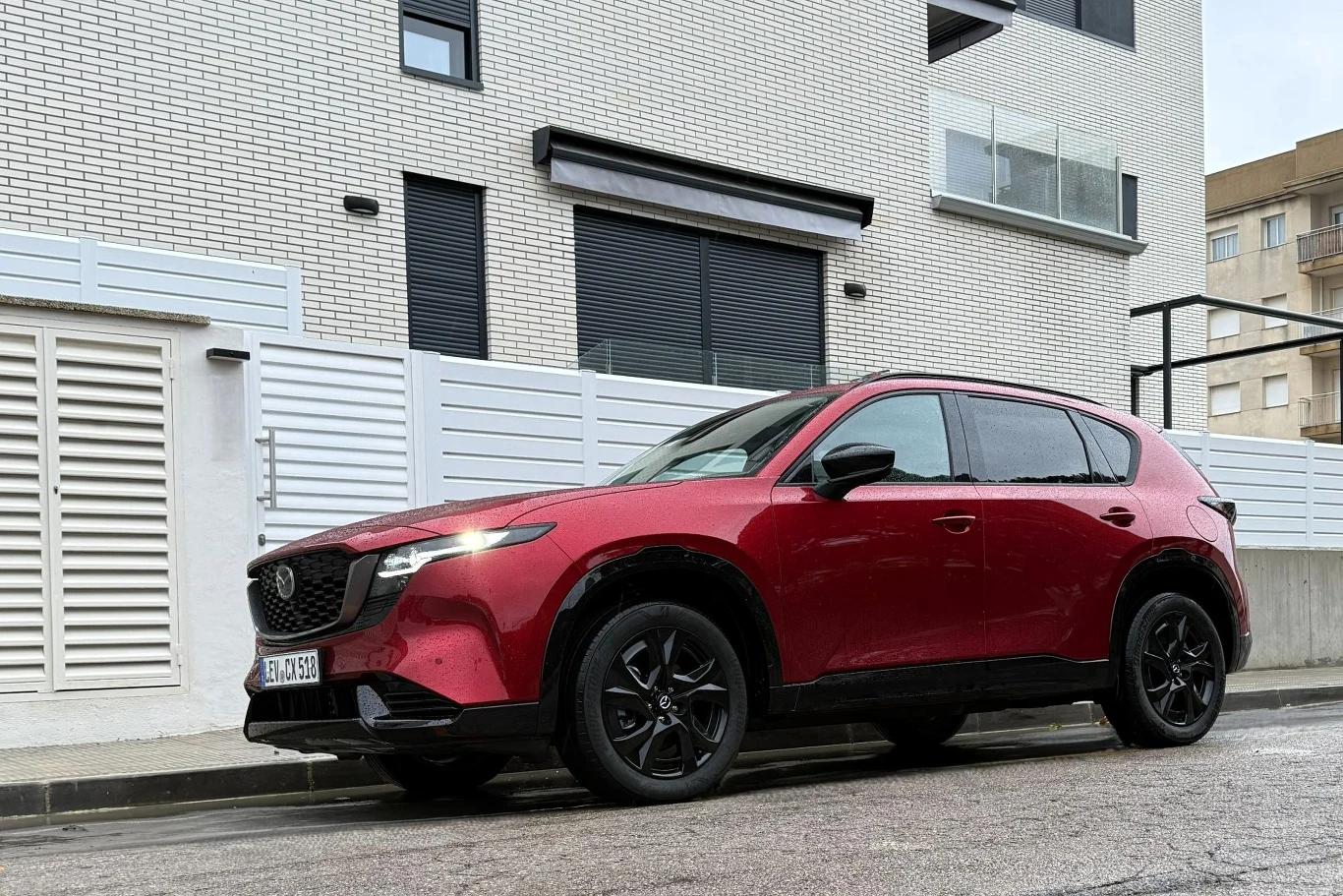 Czerwony SUV Mazda CX-5 zaparkowany przy krawężniku na tle nowoczesnego budynku mieszkalnego o jasnej elewacji i czarnych oknach.