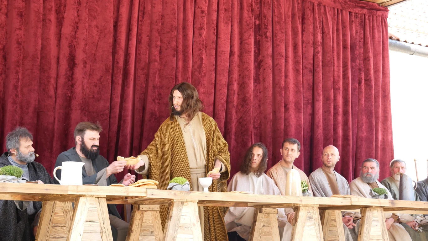 Grupa mężczyzn siedzi przy długim stole, ubranych w stroje przypominające czasy biblijne; centralna postać w brązowo-żółtym płaszczu podaje chleb innemu mężczyźnie, tło stanowią zasłony w kolorze bordowym.