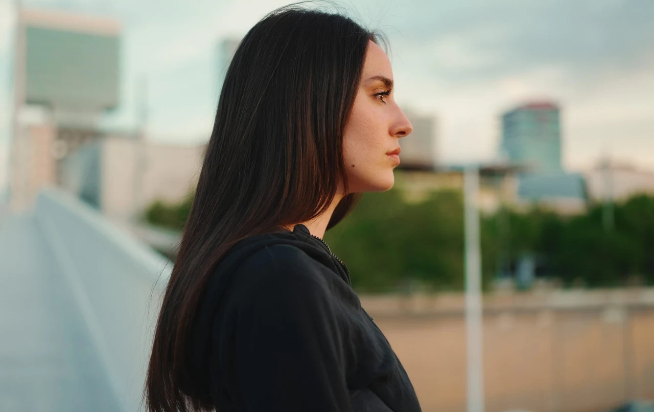 Młoda kobieta o długich, prostych, ciemnych włosach patrzy w dal, ubrana w ciemny sweter, na tle nowoczesnych budynków oraz zieleni miejskiej.