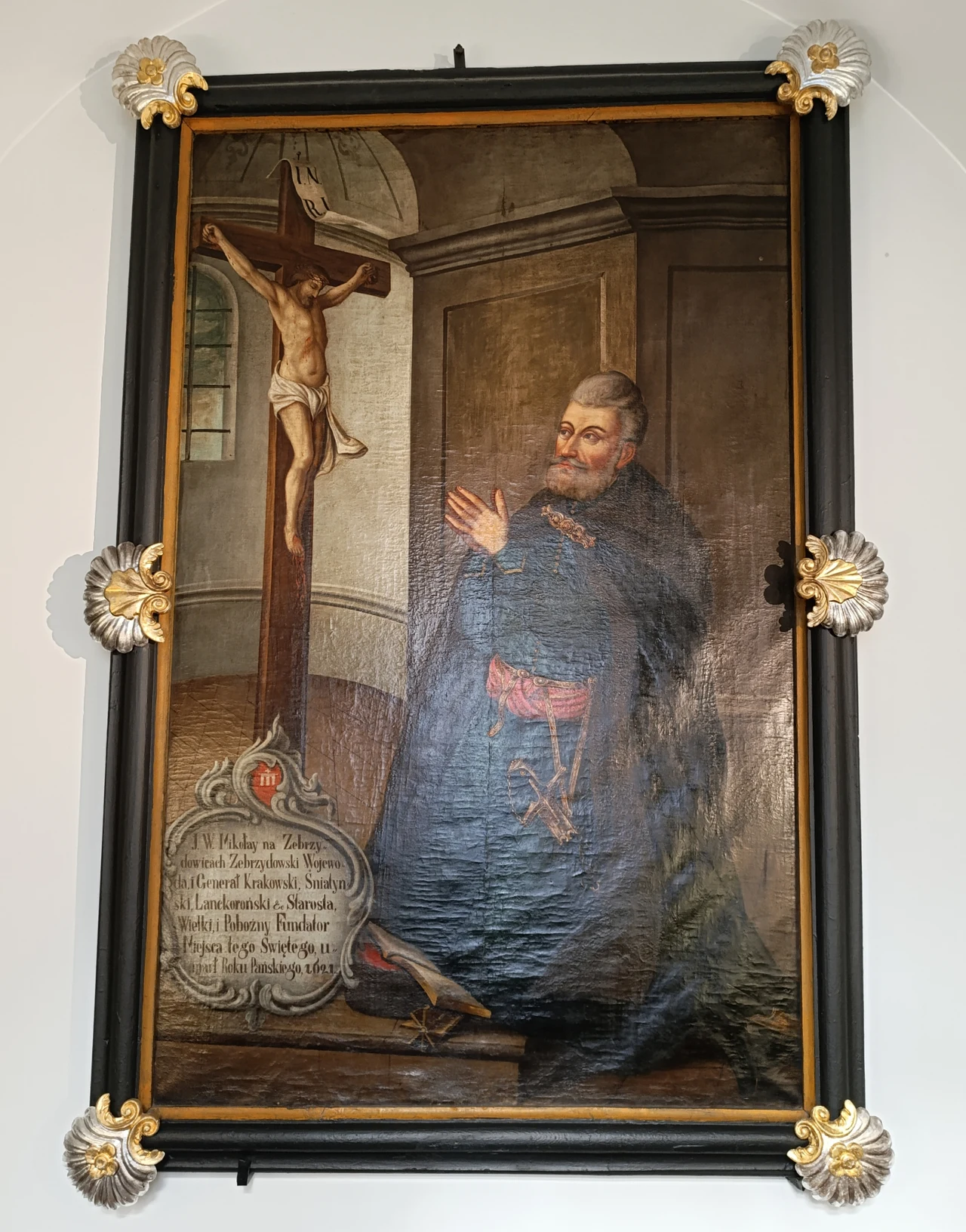 Klęczący mężczyzna w bogato zdobionym stroju szlacheckim modli się przed ukrzyżowanym Jezusem Chrystusem; na pierwszym planie widoczna inskrypcja na marmurowej tablicy, tło stanowi wnętrze z drzwiami i sklepionym sufitem.