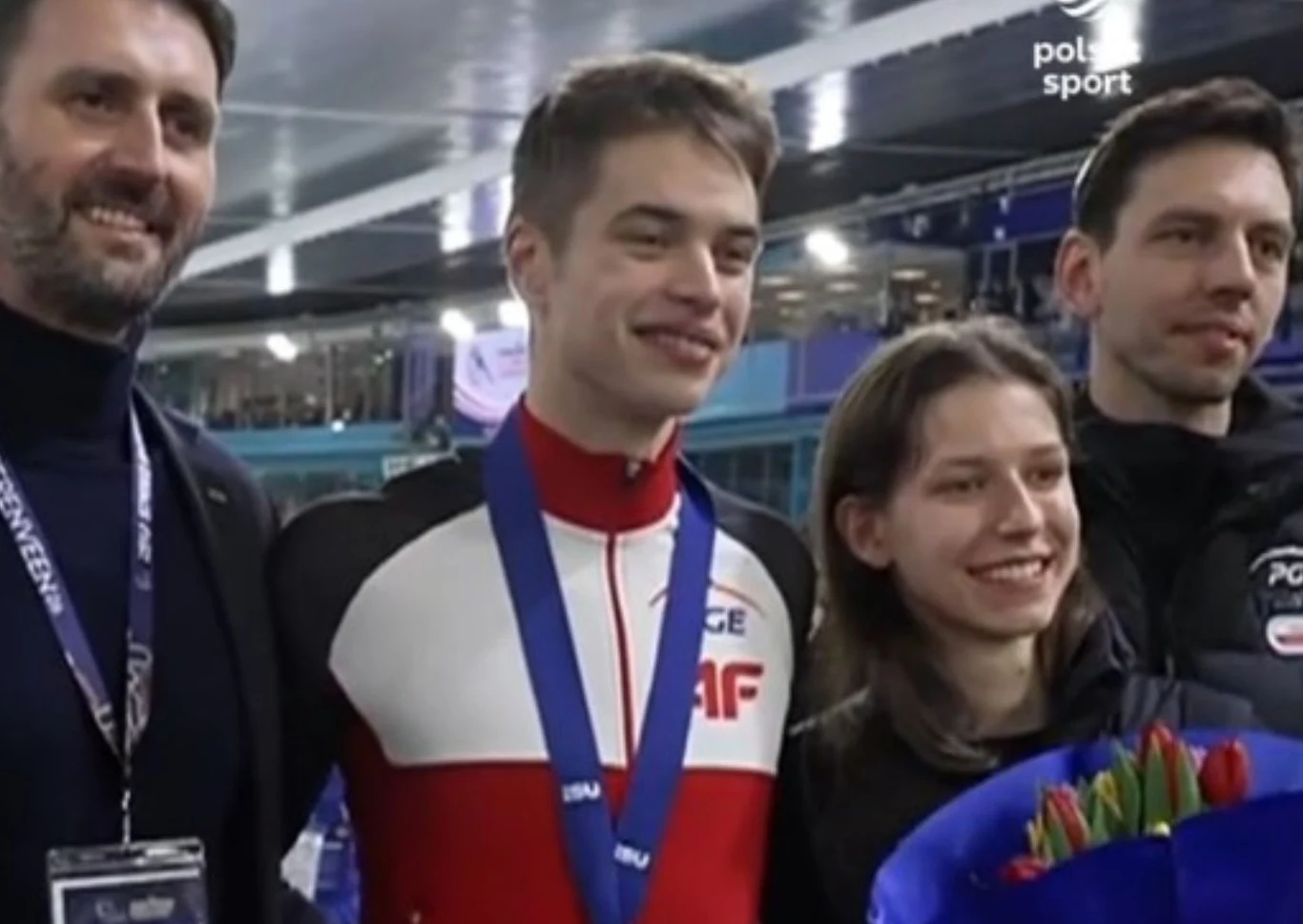 Uśmiechnięty sportowiec w biało-czerwonym stroju z medalem na szyi stoi otoczony przez trzy osoby, jedna z nich trzyma bukiet kwiatów; w tle widoczna hala sportowa.