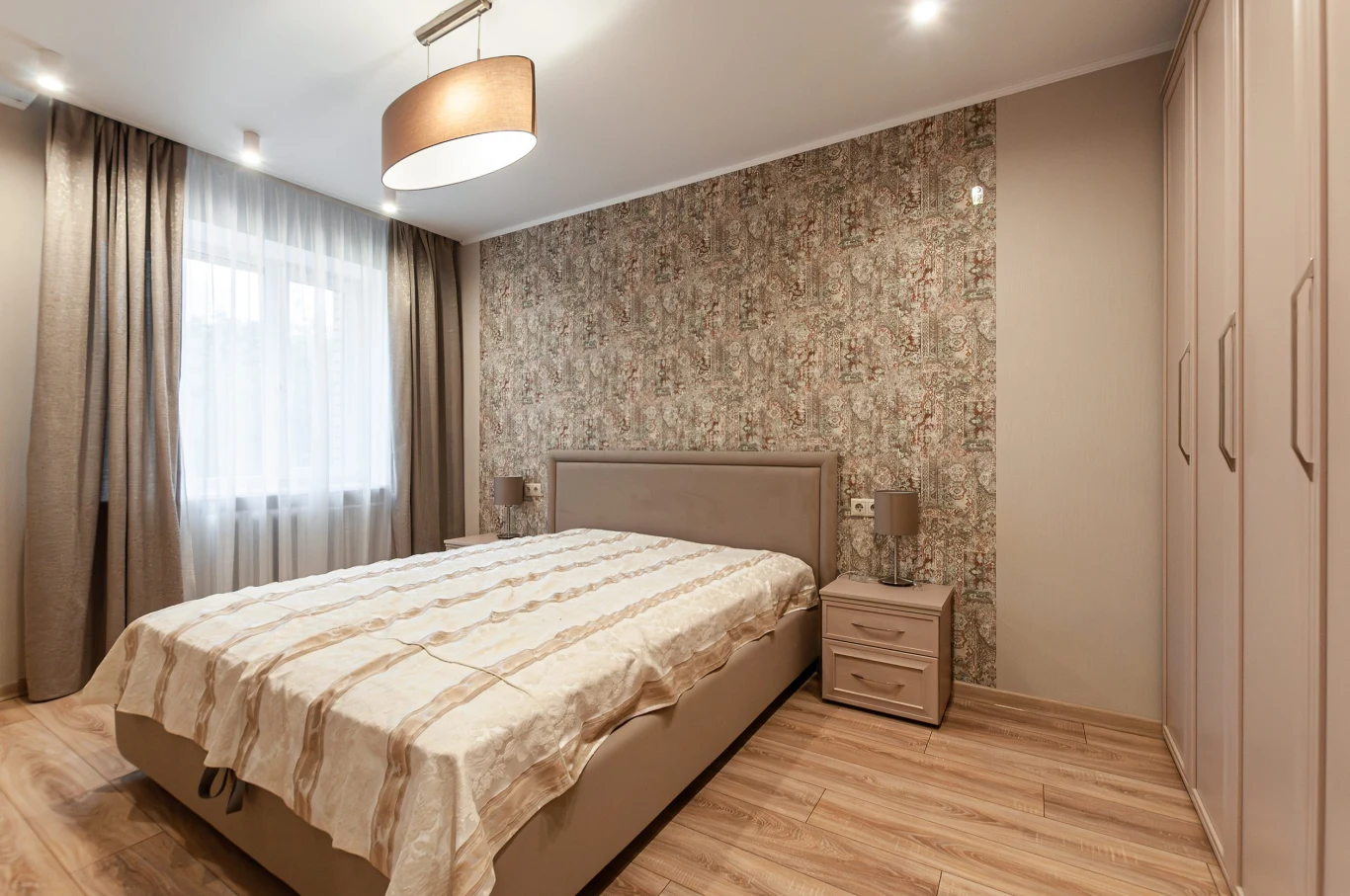 Jasna sypialnia z dużym łóżkiem na środku, nowoczesne, zabudowane szafy po prawej stronie, tapeta w delikatny wzór za wezgłowiem oraz dwa stoliki nocne z lampkami po obu stronach łóżka.