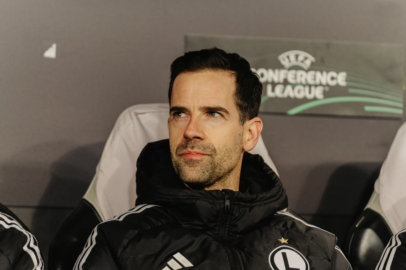 Trener w czarnej kurtce z logo Legii Warszawa siedzi na ławce rezerwowych podczas meczu Ligi Konferencji Europy, w tle widoczne logo rozgrywek.