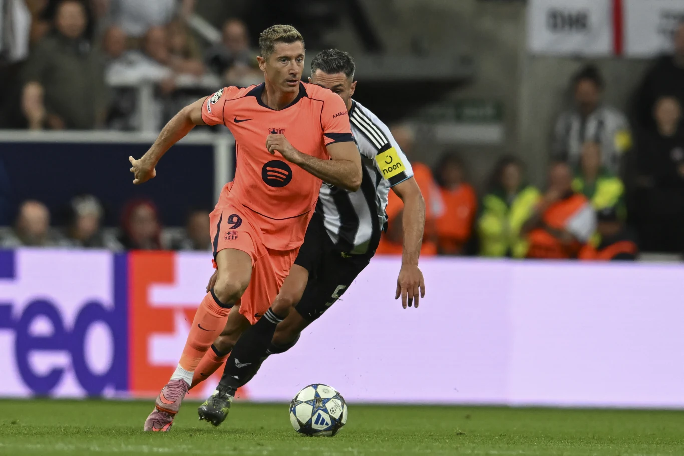 Piłkarz w różowym stroju z numerem 9 drybluje piłkę przed zawodnikiem w biało-czarnym trykocie podczas meczu piłki nożnej, wokół których na trybunach widoczni są kibice oraz stewardzi w odblaskowych kamizelkach.