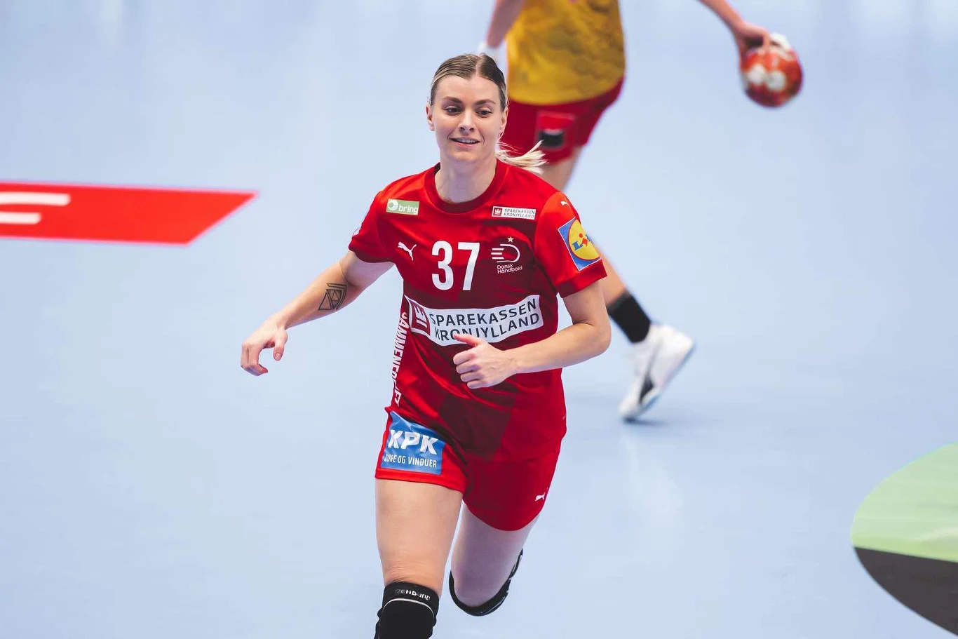 Zawodniczka w czerwonym stroju z numerem 37 grająca w piłkę ręczną na hali sportowej, dynamicznie biegnąca po parkiecie z uśmiechem na twarzy, w tle inna zawodniczka trzymająca piłkę w dłoni.