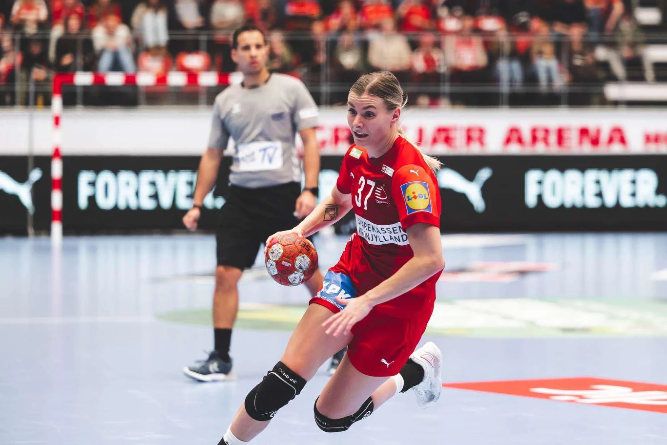 Zawodniczka w czerwonym stroju z numerem 37 dynamicznie prowadzi piłkę podczas meczu piłki ręcznej na hali sportowej, w tle widoczny sędzia oraz kibice na trybunach.