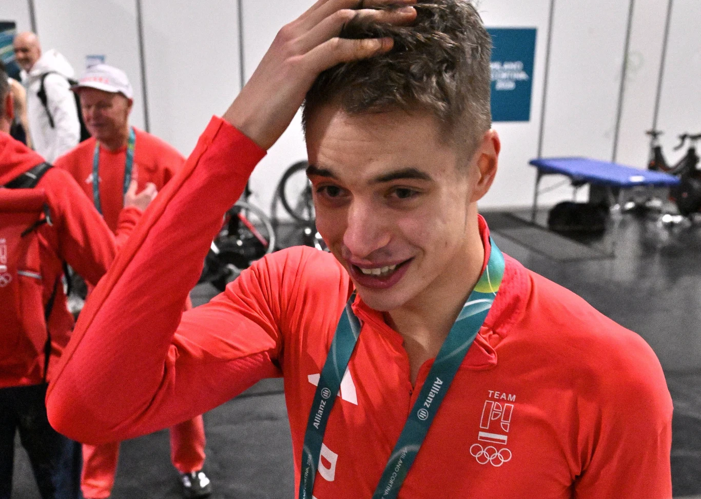 Młody sportowiec w czerwonym stroju reprezentacyjnym z biało-czerwonymi emblematami oraz medalem na szyi, z lekko zmierzwionymi włosami i uśmiechem, trzyma się za głowę w geście wyrażającym emocje; w tle inni członkowie drużyny oraz sprzęt sportowy.