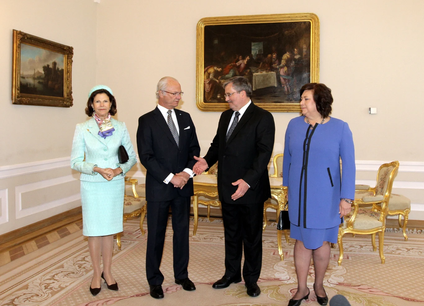 Cztery elegancko ubrane osoby stoją na tle klasycznego, gustownie umeblowanego wnętrza z bogato zdobionymi krzesłami, na ścianach wiszą dwa duże obrazy w złotych ramach, dwie osoby w centrum wymieniają uścisk dłoni.