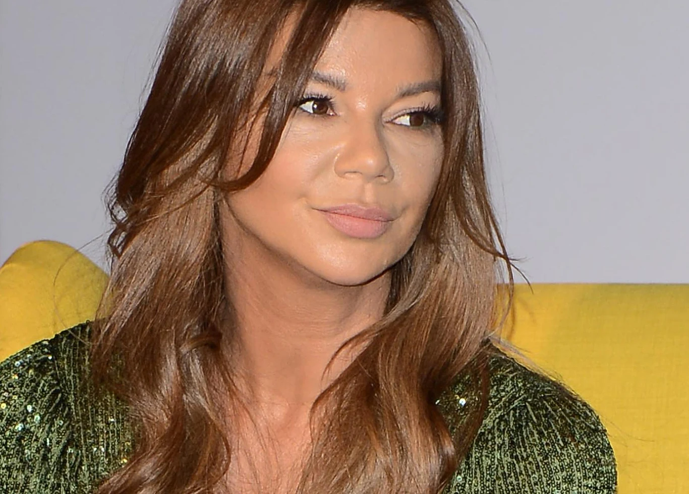 Edyta Górniak Edyta Górniak w zielonej, błyszczącej bluzce, z długimi brązowymi włosami, siedzi na żółtej kanapie.