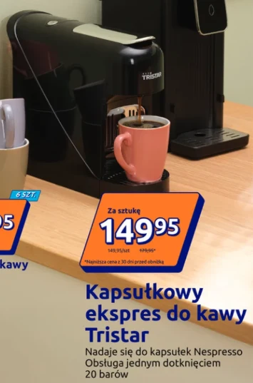 Czarny kapsułkowy ekspres do kawy marki Tristar z różowym kubkiem ustawionym na podstawce, stojący na jasnym blacie obok innego urządzenia i pojemnika z łyżeczkami; w prawym dolnym rogu widoczna jest informacja o cenie i funkcjach ekspresu.