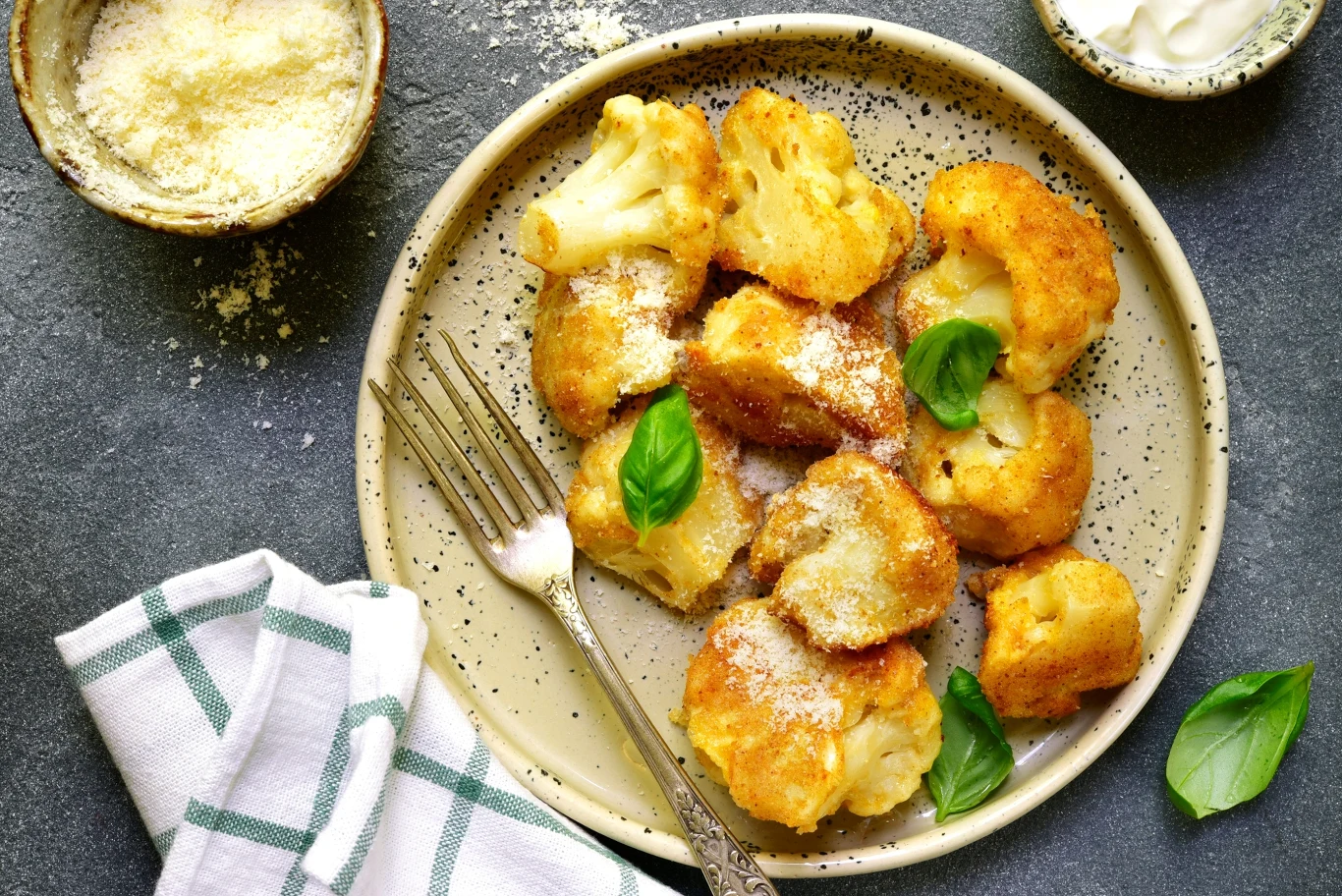 Złociste kawałki panierowanego kalafiora posypane startym serem, udekorowane świeżymi listkami bazylii, obok widelca na kremowym talerzu z ciemnymi kropkami; w tle miseczki z tartym serem i sosem oraz bawełniana serwetka.