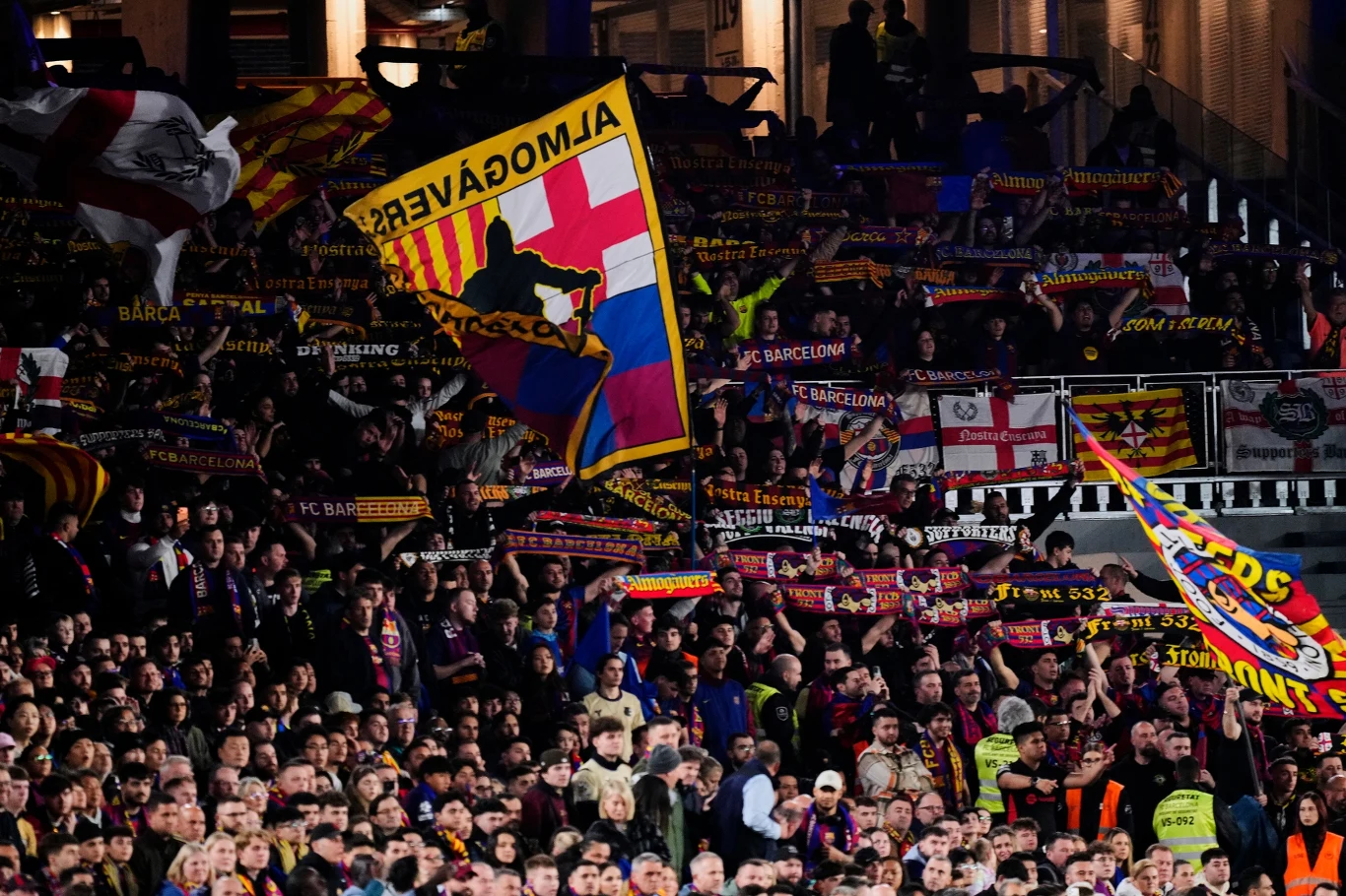 Kibice na Spotify Camp Nou Tłum kibiców FC Barcelony z kolorowymi flagami, transparentami i szalikami w barwach klubowych, w atmosferze sportowego wydarzenia, widoczni ochroniarze na przodzie tłumu.