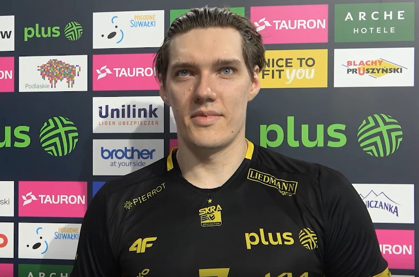 Mężczyzna w sportowej koszulce z logo Plus i innymi sponsorami stoi na tle ściany z tablicą sponsorską po zakończonym wydarzeniu sportowym.