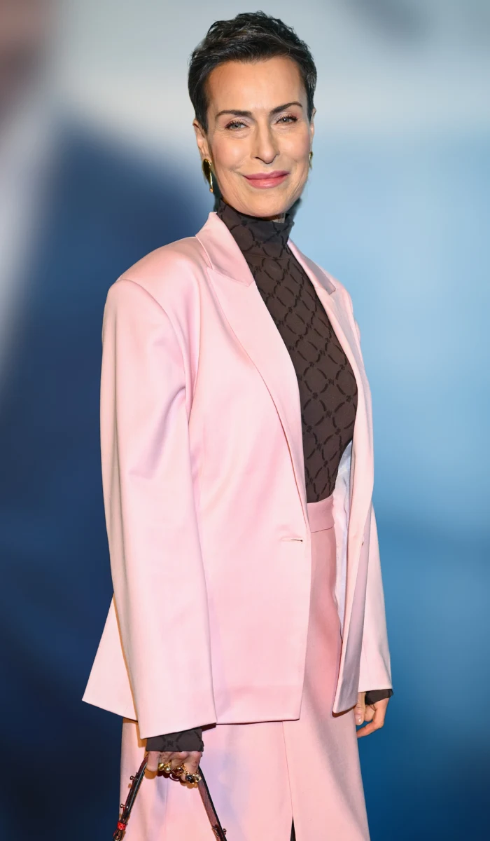 Kobieta w eleganckim, jasnoróżowym garniturze z dużą marynarką i spódnicą, pod którymi ma brązowy golf z geometrycznym wzorem; trzyma w ręku stylową torebkę na tle rozmytego niebieskiego tła.