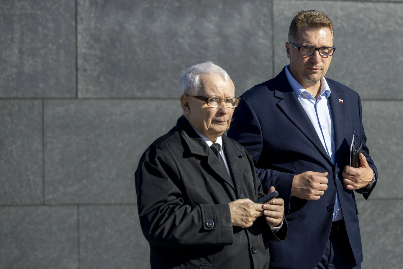 Dwóch mężczyzn w garniturach idzie obok siebie na tle szarej, betonowej ściany; starszy z nich trzyma coś w dłoniach, młodszy ma okulary i przypiętą polską flagę do klapy marynarki.