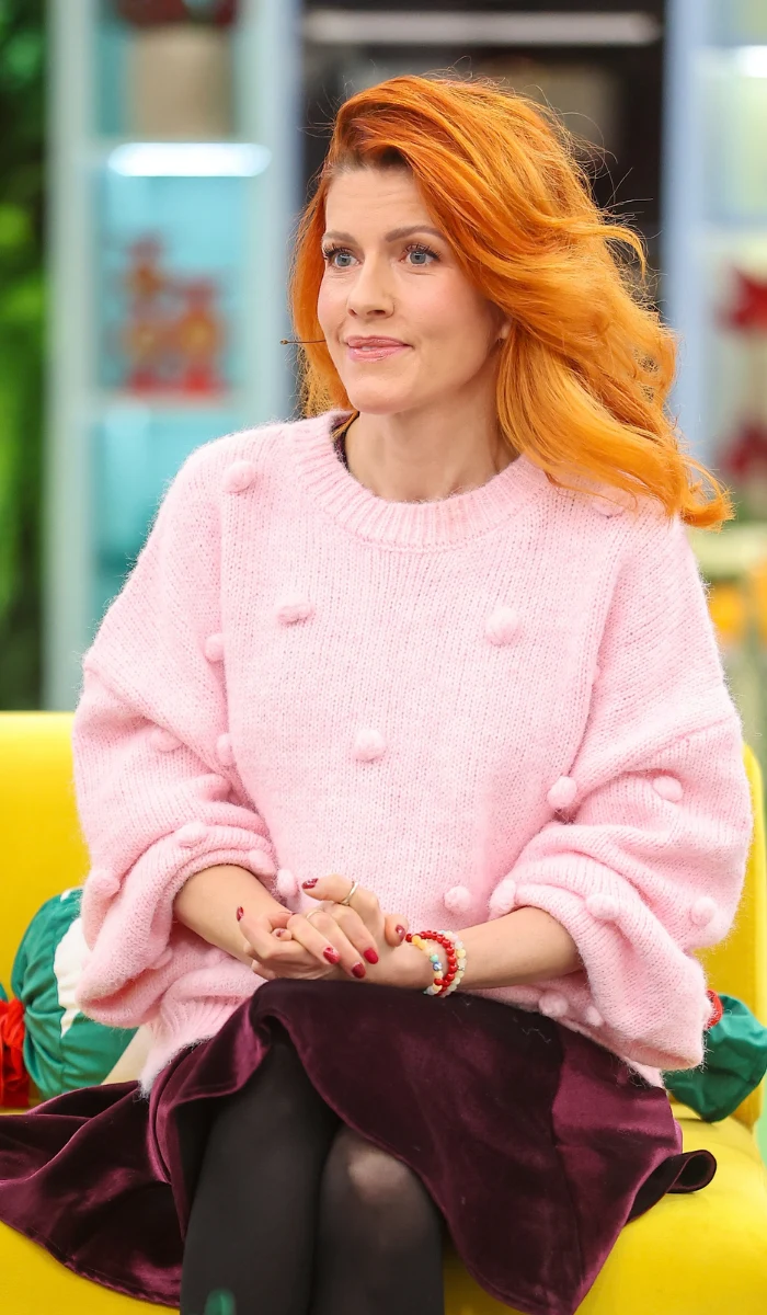 Kobieta z długimi, rudymi włosami ubrana w różowy sweter z fakturowanymi guzikami i ciemną spódnicę, siedzi na żółtej sofie z rękami złożonymi na kolanach, w tle jasne, domowe wnętrze.