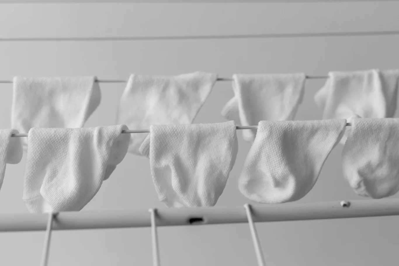 Białe dziecięce skarpetki rozwieszone na suszarce, równomiernie rozmieszczone na kilku prętach, w tle jasna, minimalistyczna przestrzeń.