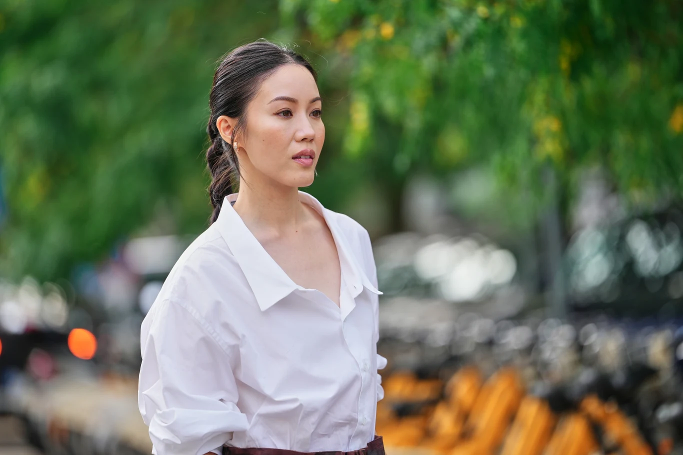 Kobieta o ciemnych włosach spiętych w niski kucyk, ubrana w białą koszulę z szerokim kołnierzem, spaceruje wśród rozmytych żółtych rowerów i zielonych drzew w tle.