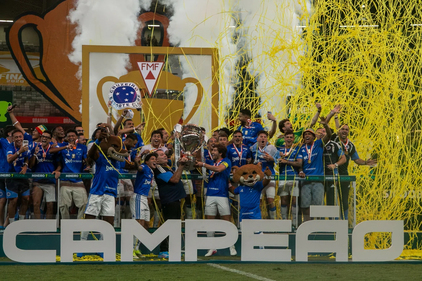 Zespół piłkarski w niebieskich strojach świętuje zdobycie trofeum, otoczony żółtymi serpentynami i dymem, za nimi znajduje się duży napis 'CAMPEAO' oraz maskotki drużyny.