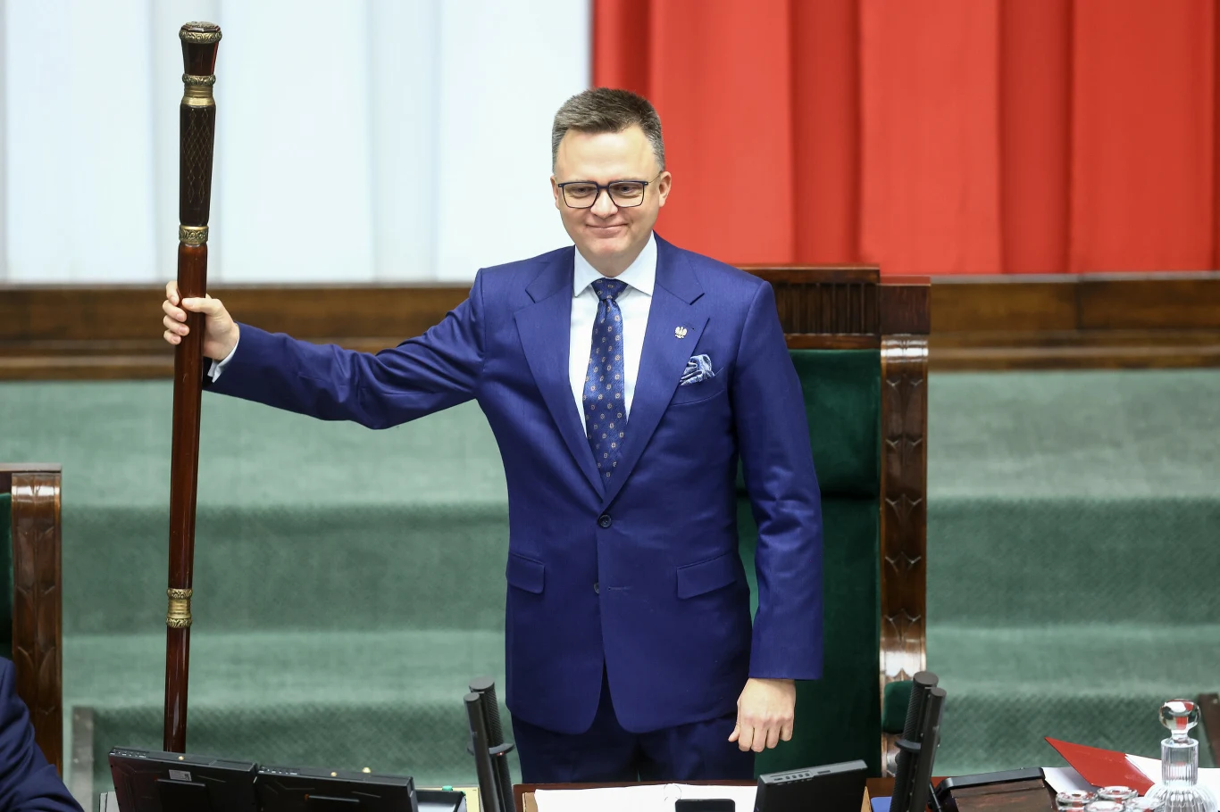 Mężczyzna w eleganckim granatowym garniturze trzyma drewnianą laskę marszałkowską, stojąc przed zielonym pulpitem w sali z wyraźnie widocznymi biało-czerwonymi pasami w tle.