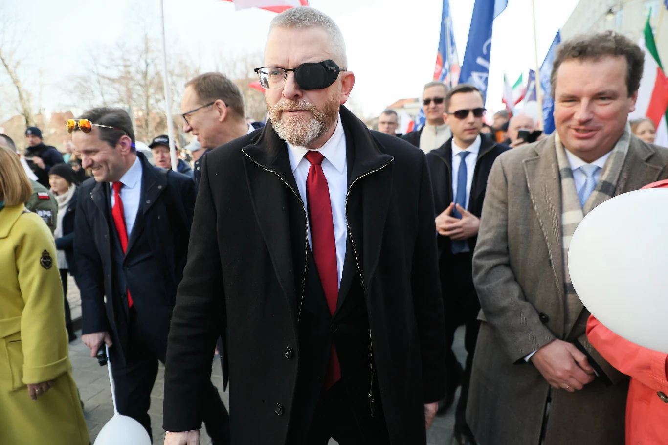 Mężczyzna z opaską na oku w czarnym płaszczu i czerwonym krawacie idzie wśród ludzi ubranych elegancko, część z nich trzyma balony i flagi, atmosfera przypomina oficjalne wydarzenie lub marsz.