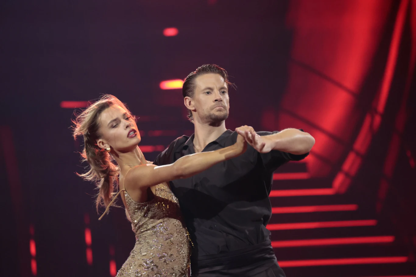 Para tancerzy w eleganckich strojach wykonuje dynamiczną pozę taneczną na scenie oświetlonej czerwonym światłem, w tle widoczne schody i elementy scenografii podkreślające atmosferę występu.