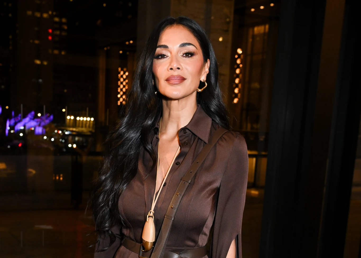 Kobieta o długich, ciemnych włosach w eleganckiej, brązowej sukience i złotych kolczykach stoi we wnętrzu z dużymi oknami na tle nocnego miasta z jasnymi światłami.