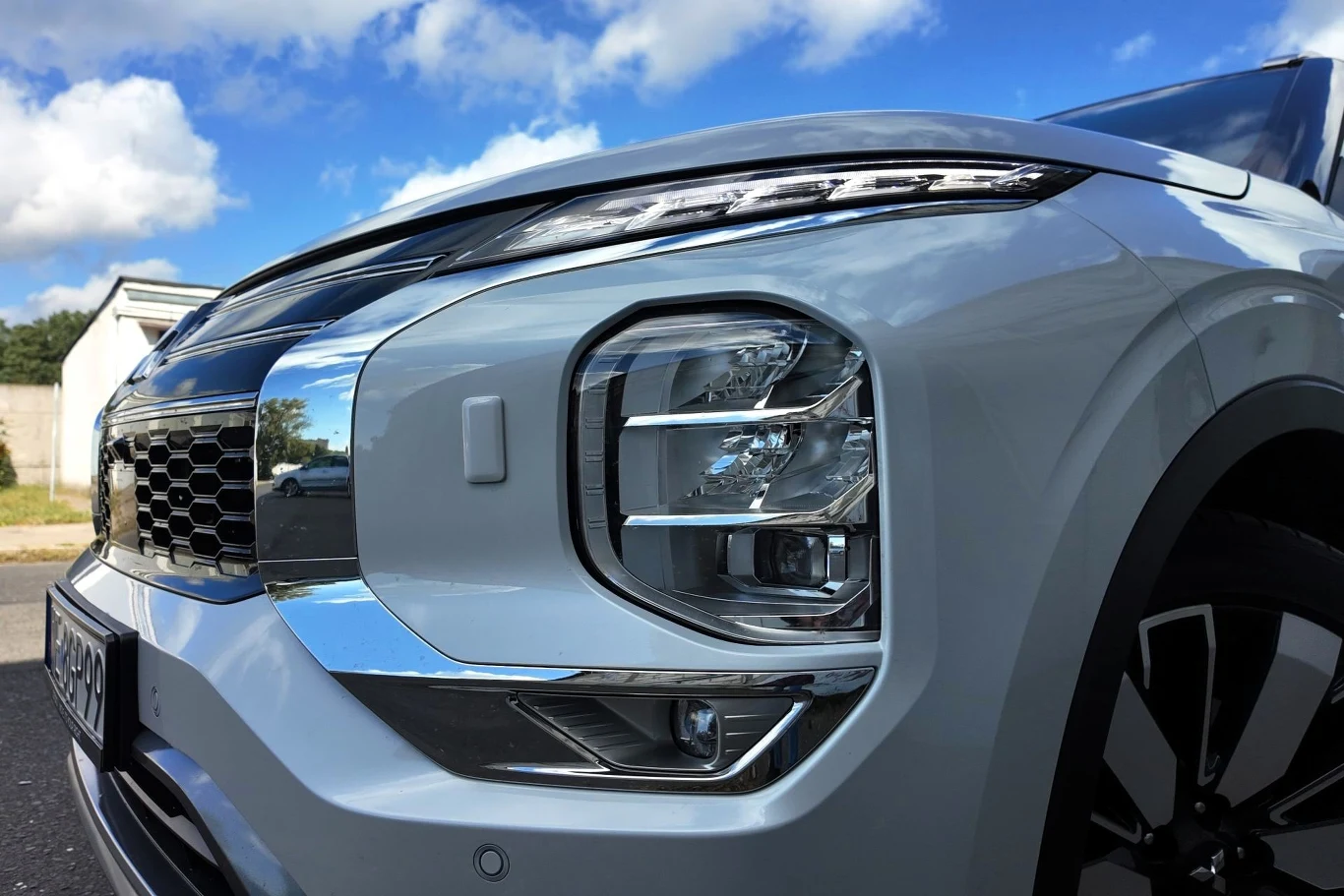 Nowoczesny przód srebrnego samochodu z dużymi reflektorami LED, chromowanymi elementami i dynamicznymi liniami maski na tle niebieskiego nieba z białymi chmurami.