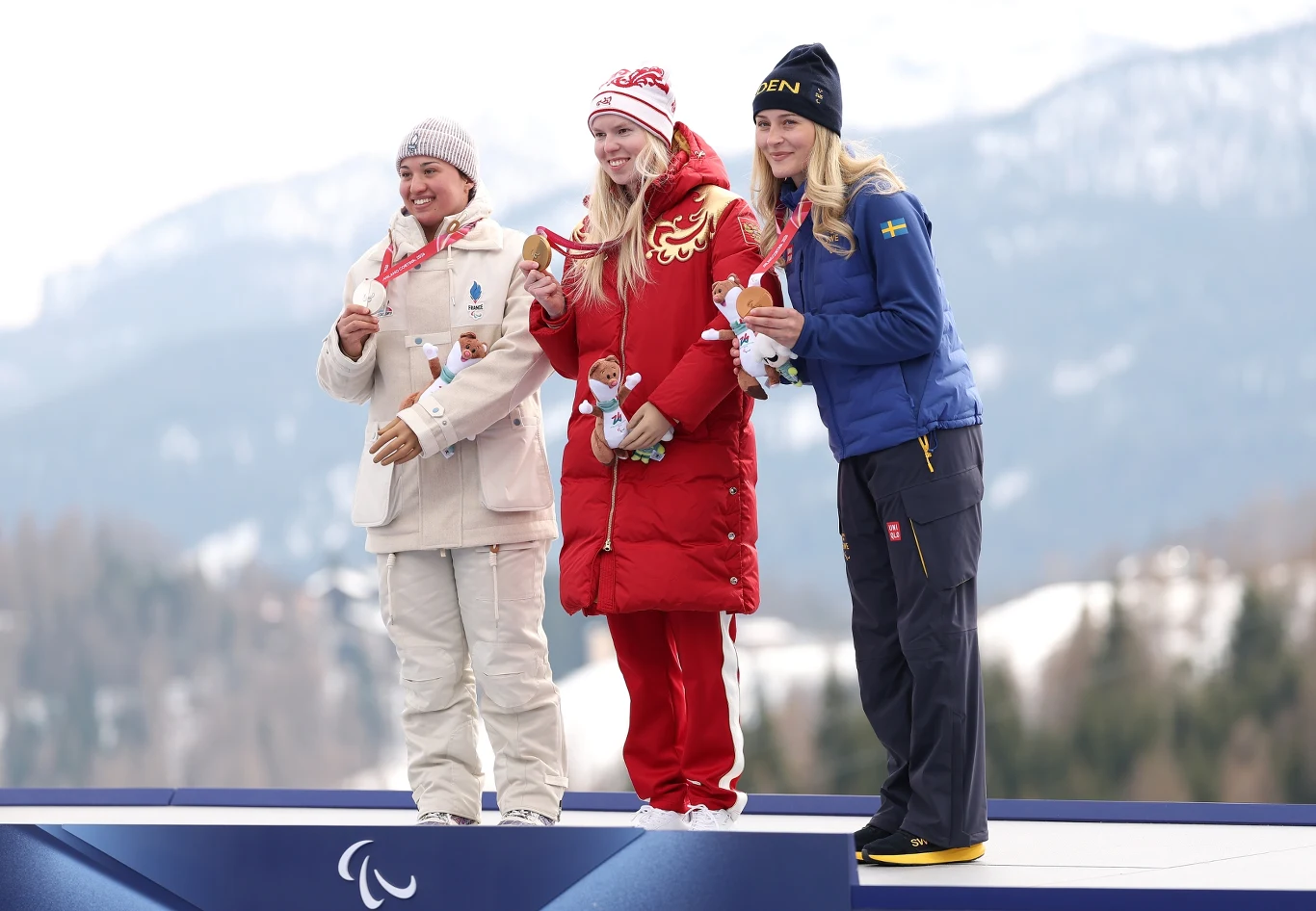 Trzy kobiety stoją na podium olimpijskim, każda z medalem na szyi; środkowa zawodniczka w czerwonym stroju z dumą trzyma złoty medal, towarzyszą jej sportsmenki w białym oraz niebieskim ubraniu, w tle zimowy krajobraz górski.
