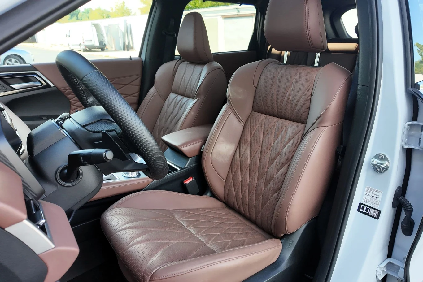 Luksusowe, brązowe, pikowane fotele samochodowe z wyraźnymi przeszyciami, nowoczesna deska rozdzielcza z dużym wyświetlaczem oraz skórzana kierownica, całość wnętrza utrzymana w eleganckim stylu.