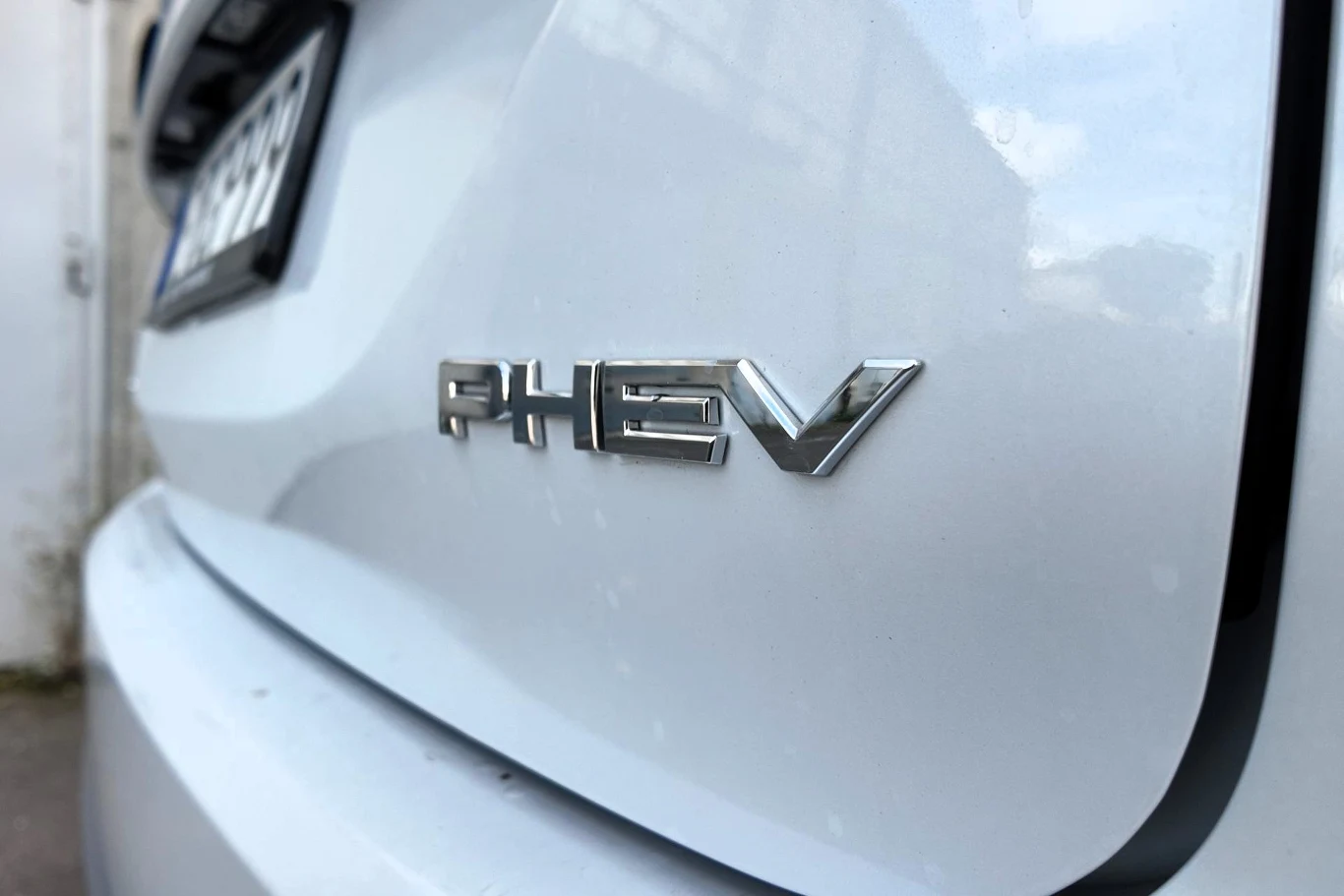 Srebrny napis PHEV umieszczony na błyszczącej, białej karoserii tylnej części samochodu, z fragmentem tablicy rejestracyjnej widocznej w tle po lewej stronie.