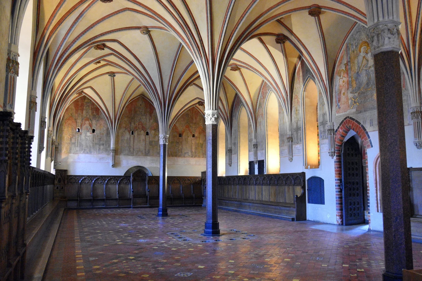 Gotycka sala z wysokim sklepieniem wspartym na kolumnach, bogato zdobionymi żebrowaniami i fragmentami polichromii na ścianach, po bokach rzędy drewnianych ław oraz widoczny portal z cegły prowadzący do kolejnego pomieszczenia.