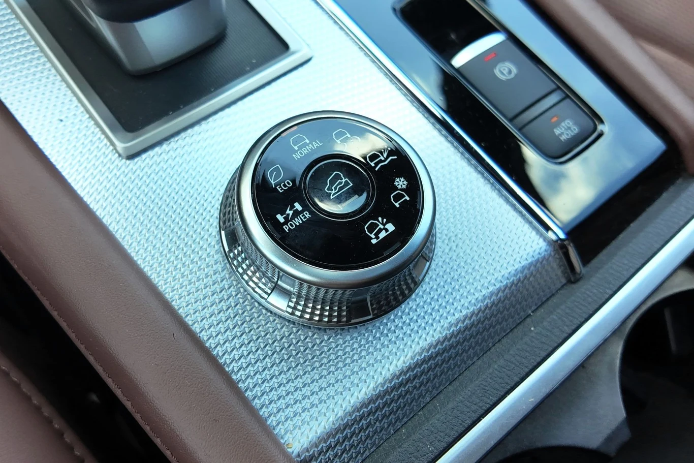 Obrotowe pokrętło do wyboru trybów jazdy w samochodzie, otoczone przyciskami kontrolnymi na metalicznej konsoli środkowej, z widocznymi detalami tapicerki i elementami wyposażenia wnętrza pojazdu.