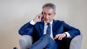 Elegancki mężczyzna w niebieskim garniturze siedzi wygodnie w szarym fotelu i rozmawia przez telefon, z lekkim uśmiechem na twarzy.