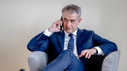 Elegancki mężczyzna w niebieskim garniturze siedzi wygodnie w szarym fotelu i rozmawia przez telefon, z lekkim uśmiechem na twarzy.