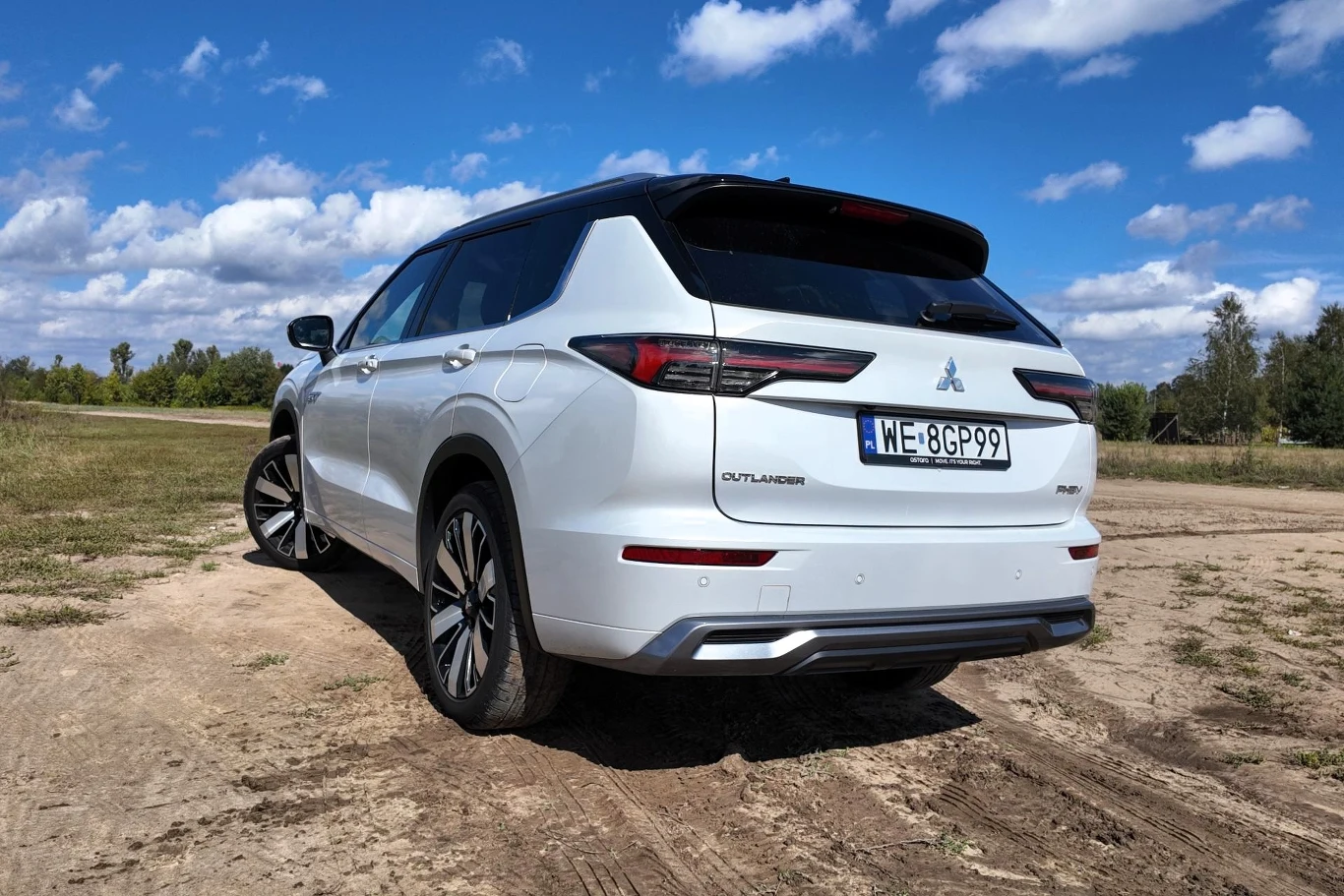 Biały SUV Mitsubishi Outlander na dużych felgach, zaparkowany na polnej drodze, otoczony przez trawiasty teren i drzewa w oddali, pod niebieskim niebem z rozproszonymi chmurami.