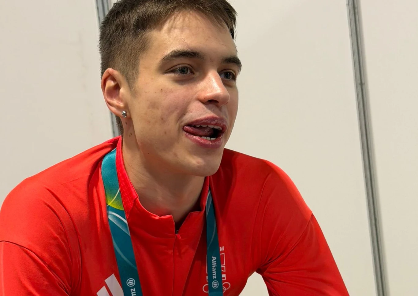Młody mężczyzna w czerwonym stroju sportowym z identyfikatorem na szyi, siedzący w jasnym wnętrzu, z lekko wysuniętym językiem i uśmiechem na twarzy.