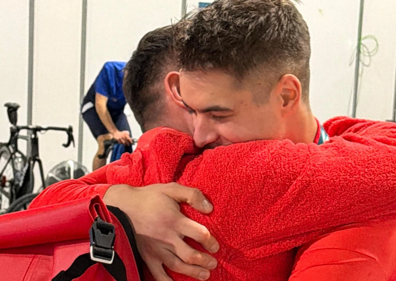 Dwóch mężczyzn w czerwonych bluzach obejmuje się serdecznie, wyrażając silne emocje; w tle widoczny rower i osoba w niebieskim stroju.