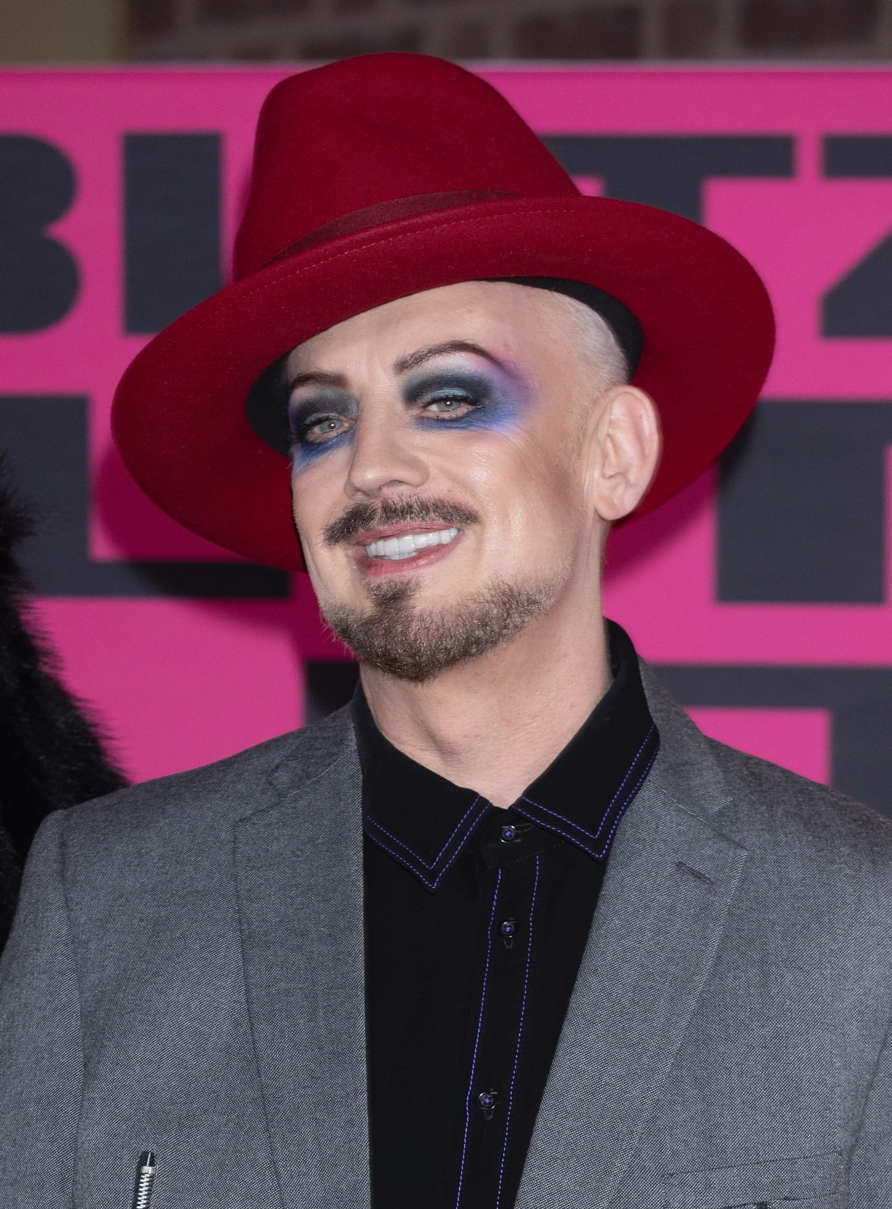 Mężczyzna w jasnoczerwonym kapeluszu i szarej marynarce, z wyrazistym makijażem oczu w odcieniach niebieskiego i fioletu, uśmiechnięty na tle różowej ściany z czarnymi literami.