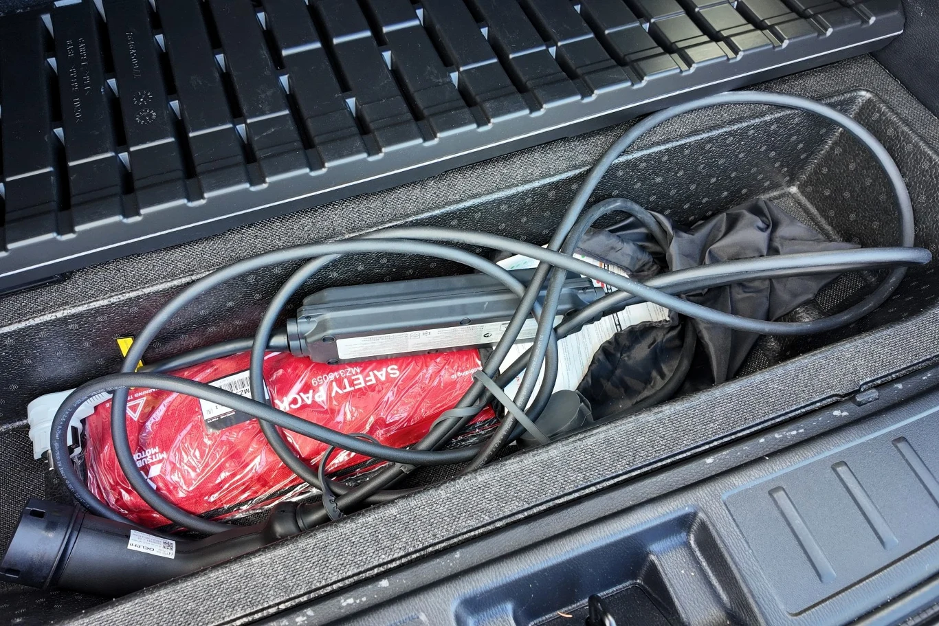 Schowek bagażnika samochodowego z przewodem do ładowania pojazdu elektrycznego, czerwoną torbą oznaczoną jako 'safety pack' oraz złożonym czarnym parasolem.