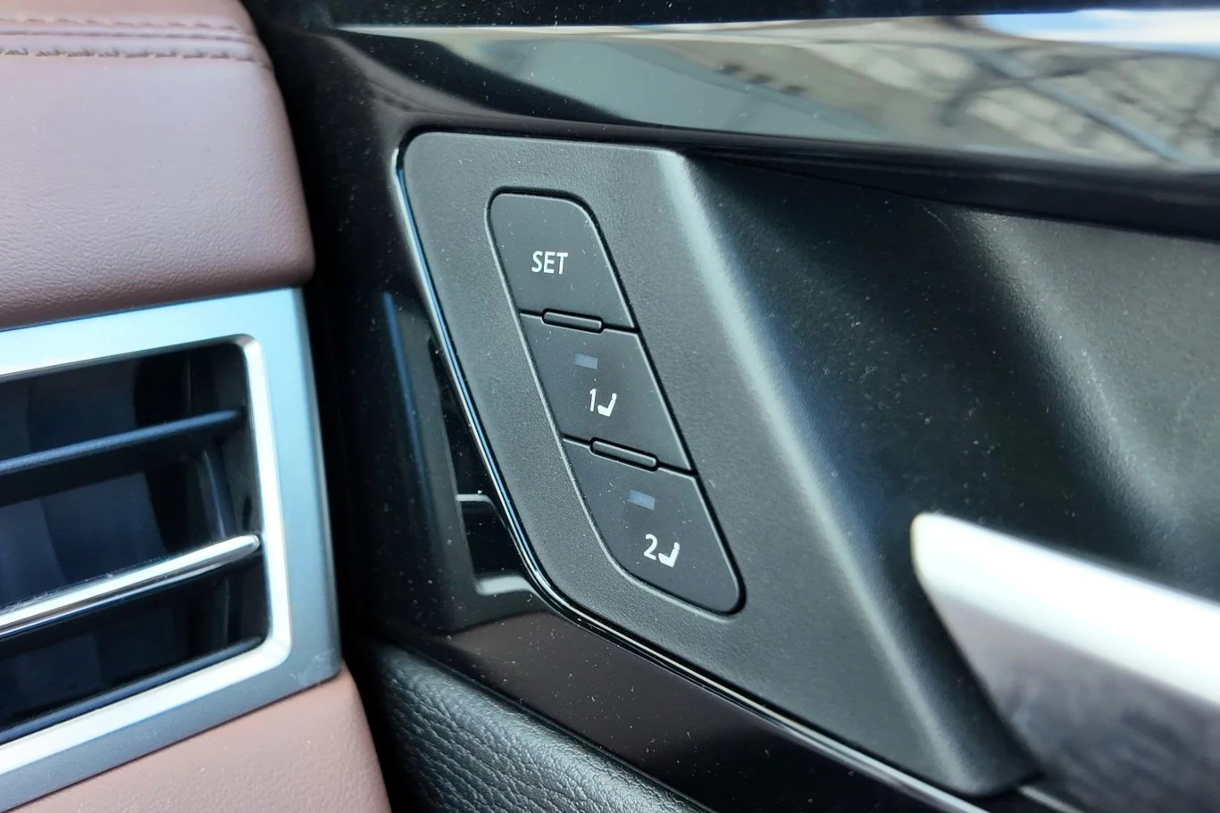 Panel przycisków do ustawiania pamięci pozycji fotela kierowcy w samochodzie, z trzema wyraźnie oznaczonymi przyciskami: SET, 1J i 2J, otoczony elementami wnętrza pojazdu w kolorze czarnym i brązowym.