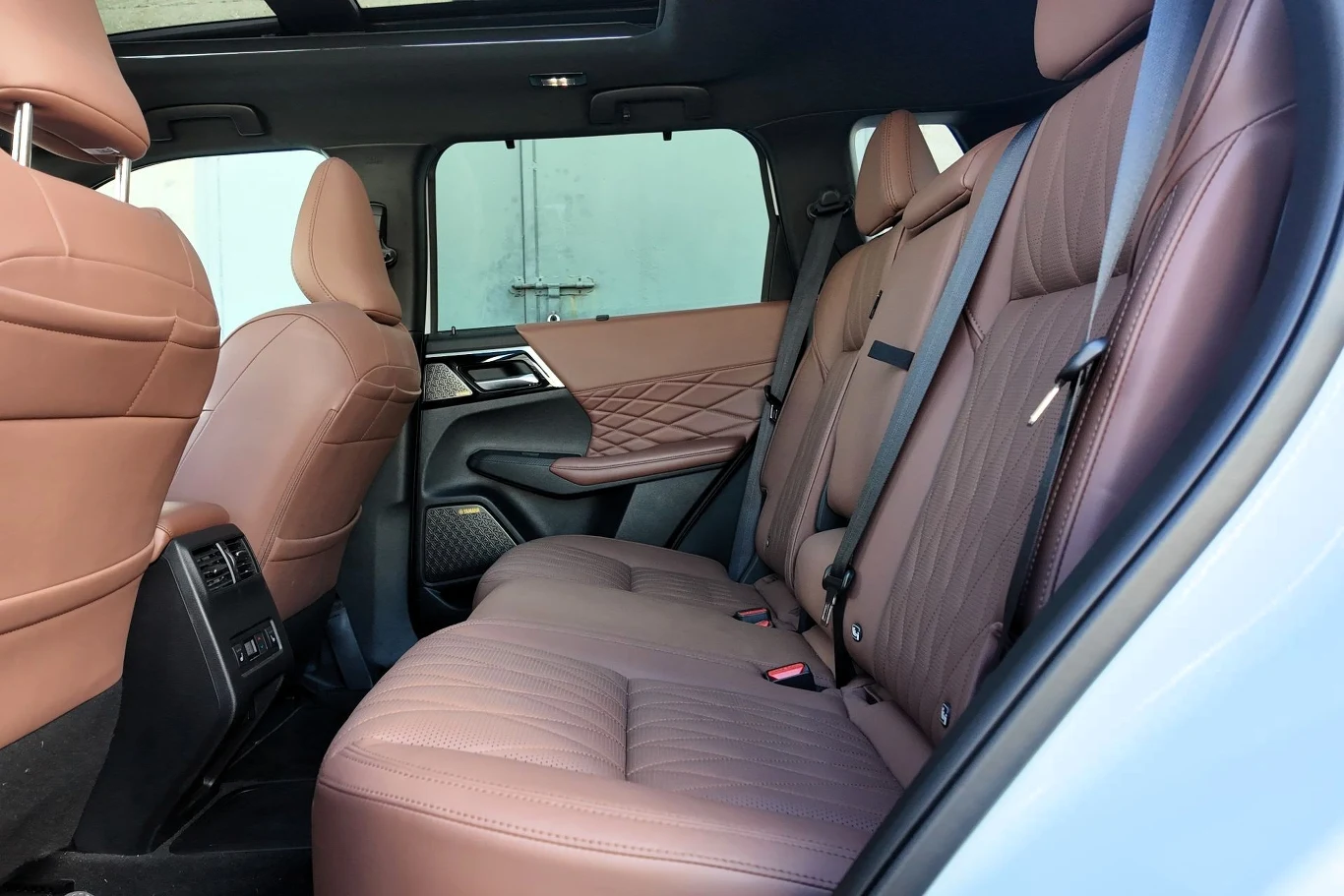 Brązowe, skórzane siedzenia tylnej kanapy samochodu z widocznymi przeszyciami, eleganckimi zagłówkami i pasami bezpieczeństwa, wnętrze utrzymane w nowoczesnym stylu z wyraźnie zaznaczonymi detalami tapicerki drzwi.