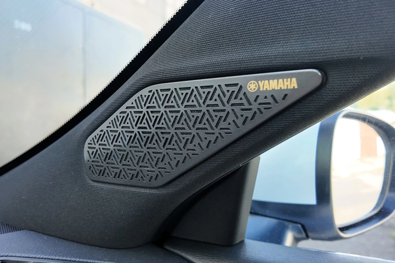 Czarny głośnik z geometrycznym wzorem i złotym logo Yamaha zamontowany w słupku samochodowym obok bocznego lusterka.
