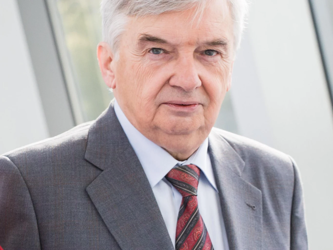 Tadeusz Sznuk Tadeusz Sznuk w szarym garniturze, białej koszuli i krawacie, stoi przy dużych oknach w nowoczesnym wnętrzu.