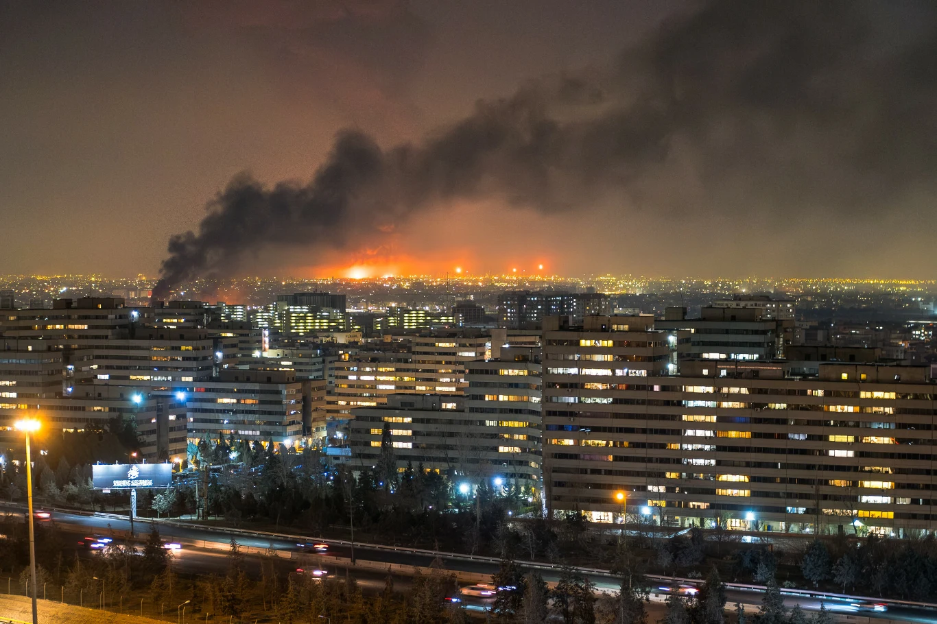 Nocna panorama miasta z gęstą chmurą czarnego dymu unoszącą się nad jednym z budynków, świadomość pożaru widoczna przez intensywne światło ognia oraz oświetlone okna bloków mieszkalnych na pierwszym planie.