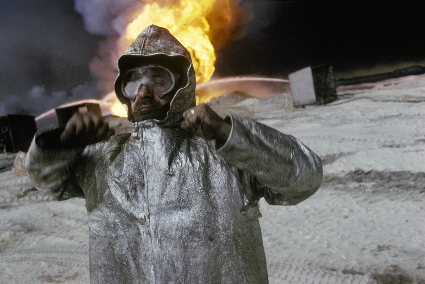 Człowiek w odbijającym światło kombinezonie ochronnym, z goglami na twarzy, stojący na tle ogromnego wybuchu ognia w przemysłowym, pokrytym śladami opon terenie.